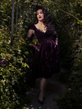 The model sits gracefully on a velvet settee, the full skirt draping around her in elegant folds.