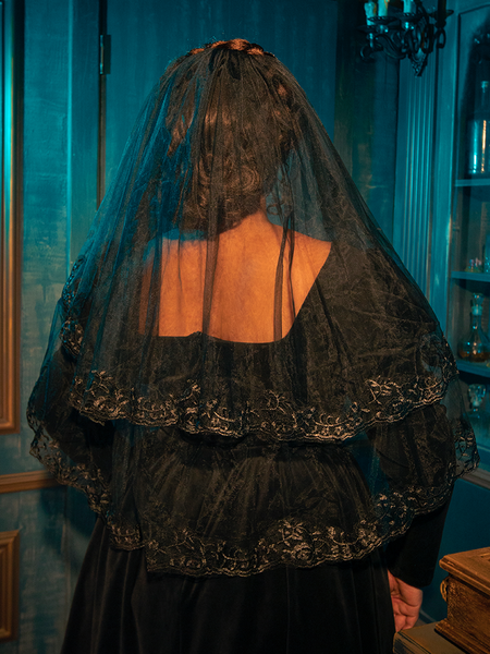 Pensive and captivating, a Gothic female model skillfully exhibits the Black Veil with Embroidered Lace Trim from La Femme en Noir, incorporating a touch of reflection into the ensemble with each striking pose.