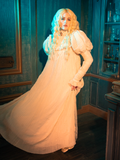 The CRIMSON PEAK™ Edith Victorian Gown glows like a moonlit sonata, its ivory fabric shimmering under the caress of the night. The intricate lace detailing and ruffled sleeves compose a melody of elegance and mystery, inviting the wearer to lose themselves in the romance of the Victorian era.