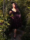 Seductively positioned amidst lush greenery, Micheline Pitt dons the Baudelaire Swing Dress in Plum from the gothic brand La Femme en Noir.