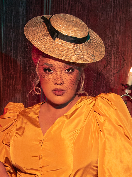 A haunting allure envelops the scene as a female gothic model gracefully showcases the Wicket Tilt Hat with Black Veil from the mysterious La Femme en Noir, embodying the dark elegance of the brand.