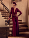 Aliza, standing on the staircase of a palatial home, models the Art Deco ruched gown in crimson from La Femme En Noir.