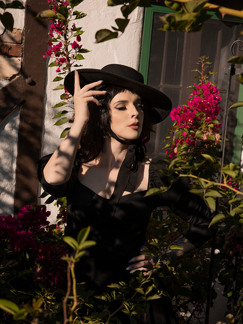 Female model wears the Bolero Hat in Black from La Femme en Noir while standing in a garden.