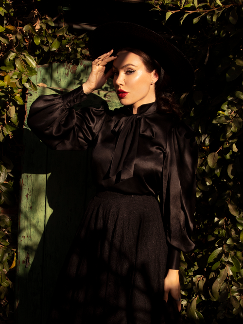 Micheline Pitt photographed in a green garden setting wearing a gothic style clothing outfit including the Victorian Blouse in Black.