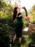 Micheline Pitt turned away from the camera to show off the back of the Baudelaire Wiggle Dress in Hunter Green While gently smelling a red rose.