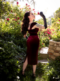 Micheline Pitt, facing away from the camera and holding a pink rose up to her nose, models a gothic style retro dress from La Femme en Noir.