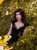 Stephanie sitting in a lush garden area wearing the goth style dress entitled the Baudelaire Wiggle Dress in Black from La Femme en Noir.