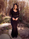 Gently lit by light, Rachel Sedory wears the Black Marilyn Lace Gown in Black while standing in a bamboo garden.