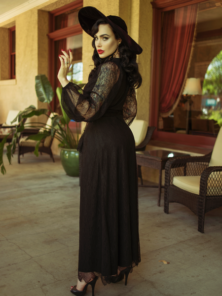 Micheline Pitt with her body facing away from the camera, subtly turns her head back while wearing the Black Widow Wrap Gown in Black Lace.