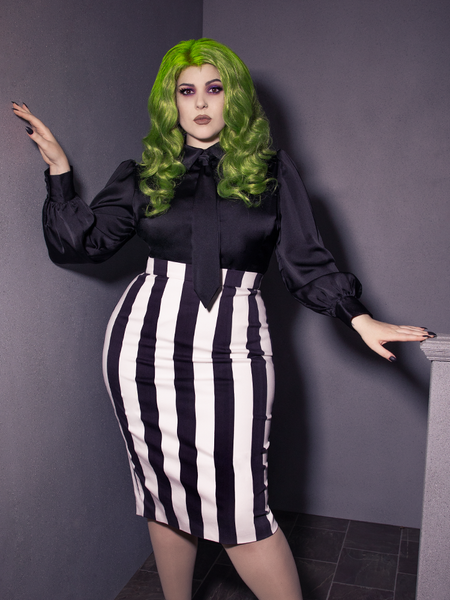 Female model standing in a grey stairwell modeling a gothic glamour outfit highlighted by the Bowie Blouse w/Matching Tie in Black Charmeuse.
