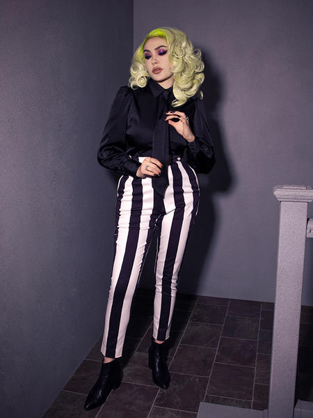 Micheline Pitt adjusting the black tie on the Bowie Blouse w/Matching Tie in Black Charmeuse she's wearing with black and white striped pants and black ankle boots. All items available from goth glamour clothing brand La Femme en Noir.