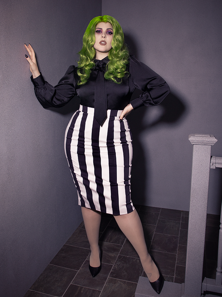 Full length image of female with green hair wearing a long sleeve black shiny top tucked into a black and white striped skirt. The outfit is finished off with black flats.