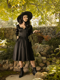 Full length shot of Stephanie standing in a lush garden while wearing the latest goth style dress from La Femme en Noir - the Cottage Witch Dress in Japanese Black Satin.