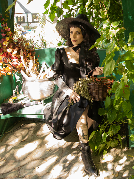 Stephanie Joens sitting on a green bench amongst many plants and trees while modeling the all-new Cottage Witch Dress in Japanese Black Satin from goth retro clothing company La Femme en Noir.