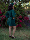A back shot of Erika, with her hands on her hips in a garden, models the Black Widow tap shorts in hunter green from La Femme En Noir.