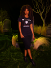 Female model standing in a graveyard scene while wearing the BEETLEJUICE™ Lydia Dead Tee with a knee-length black skirt and black ankle boots.