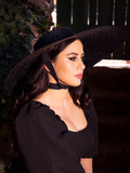 Profile shot of Ashley sitting in a garden sitting while wearing the Lydia Sun Hat from goth clothing brand La Femme en Noir.