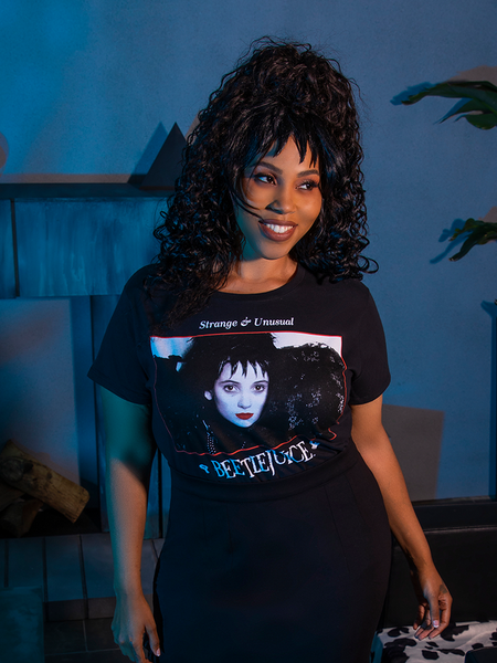 Female model smiling as she looks off camera while wearing the BEETLEJUICE™ Lydia Dead Tee tucked into a black skirt.