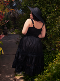 Embracing the sun's warmth in a vibrant garden, a stunning brunette model becomes a vision of elegance, showcasing the mesmerizing Pickety Witch Dress in Black, an exquisite gothic dress crafted by La Femme en Noir.