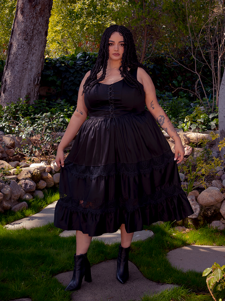 Under the golden rays of the sun, a brunette model flourishes in a vibrant garden, showcasing the striking Pickety Witch Dress in Black, a remarkable gothic dress creation by La Femme en Noir.