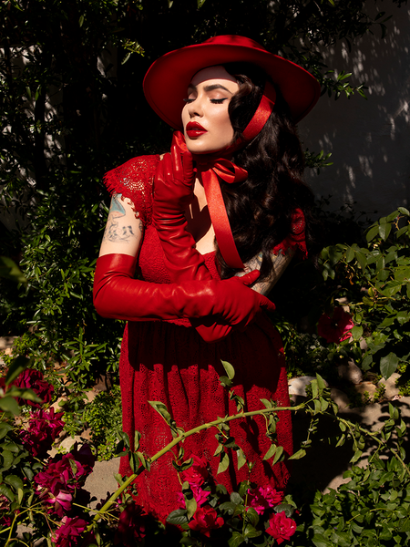 Micheline Pitt modeling the Bolero Hat in Crimson while standing in a rose garden.