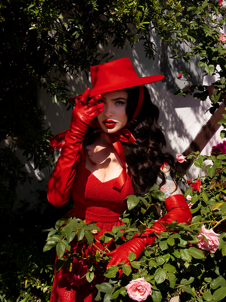 Looking at the camera, Micheline Pitt touches the brim on her Faux Leather Opera Gloves in Crimson.