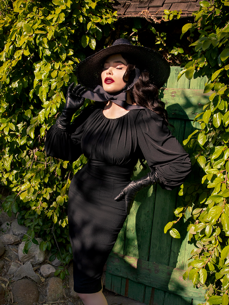 Micheline Pitt posing with her Salem Top in black paired with a black pencil skirt and long sleeve flowy top.