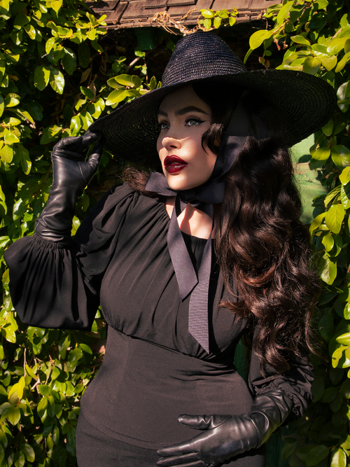 Micheline Pitt wearing an all black ensemble including the Salem Top in Black with complimentary black witch hat and vegan leather gloves.