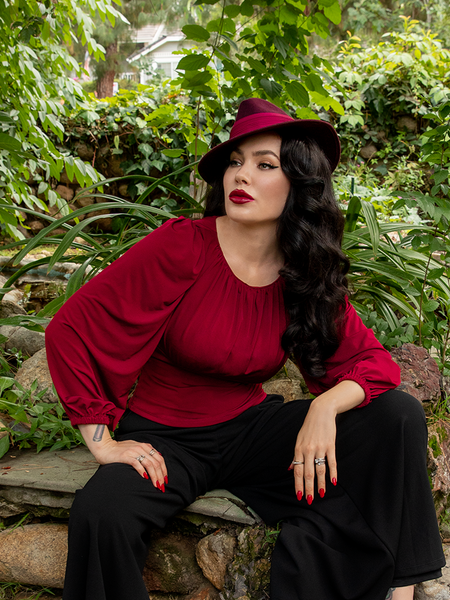 Micheline sitting in a green garden setting while modeling the Salem Top in Oxblood.
