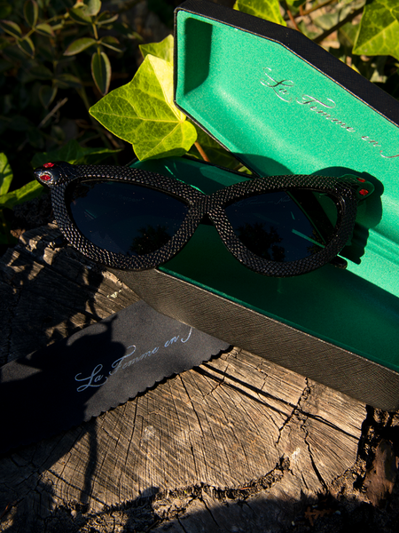 Product shot of the Serpent Sunglasses in Black sitting on top of the open coffin-shaped carrying case and next to the branded cleaning cloth.