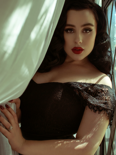 Rachel leans on a window and pulls back a white curtain while modeling the Southern Gothic bustier top in black.