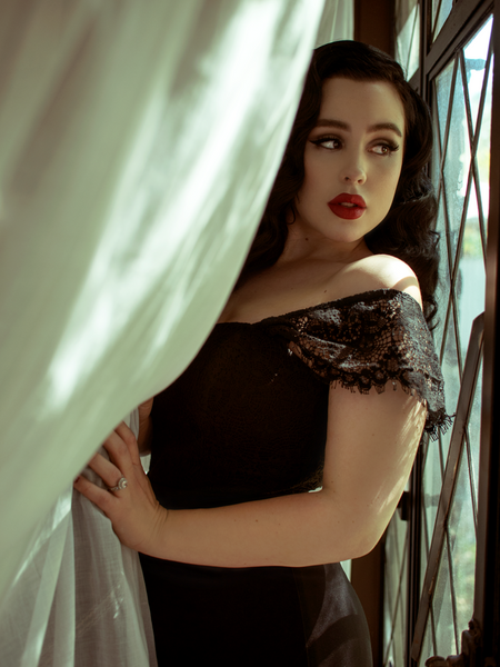 Rachel leans on a window and pulls back a white curtain while modeling the Southern Gothic bustier top in black.