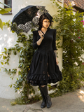 Full length shot of model wearing a black gothic style dress paired with a matching parasol and knee length black boots.