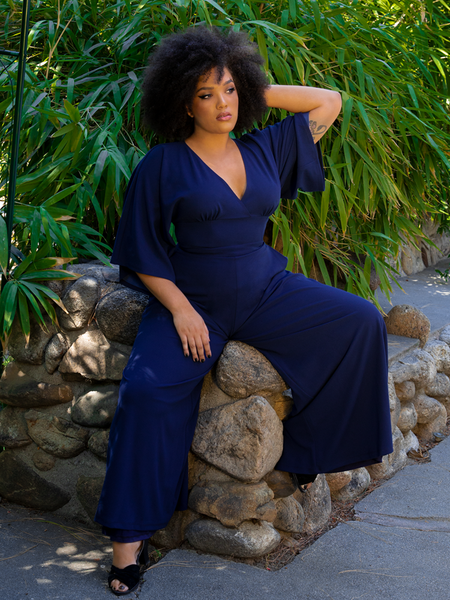 Ashleeta sitting on the arm of a stone rock bench while wearing the Black Widow Palazzo Pants in Navy from gothic retro clothing company La Femme en Noir.
