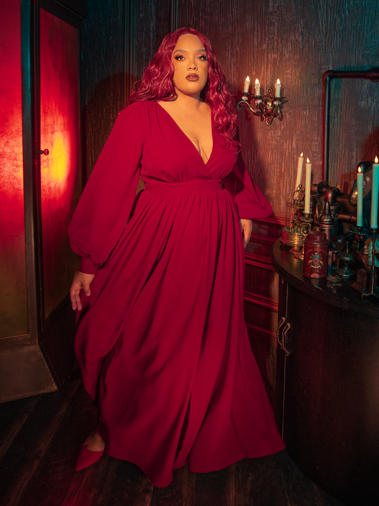 A model wearing the crimson red Belladonna maxi dress stands in a dimly lit garden, the red fabric contrasting against green foliage for a Whimsigoth and dark aesthetic.