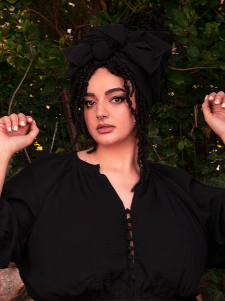 Behold the hauntingly beautiful model, adorned with gothic splendor, as she graces us with the mesmerizing Coven Head Wrap in Black, a bewitching creation from the darkly romantic realm of La Femme en Noir Clothing.