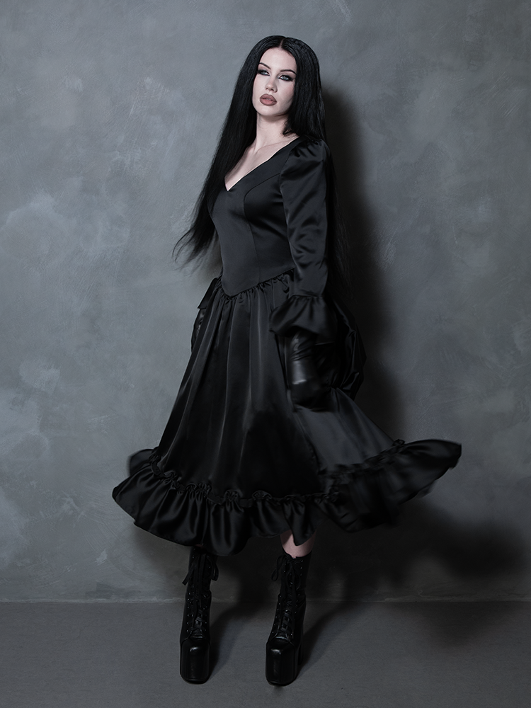 A female model highlights a dramatic Whimsigoth style vibe while wearing a flowing black satin dress rich with dark elegance.