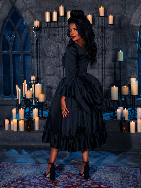 Amidst the dimly lit chamber, illuminated solely by the eerie glow of candles, the exquisite maiden unveils the Raven-Hued Romance Satin Bustle Dress from the abyssal collections of the gothic couturier, La Femme en Noir.