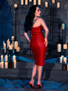 In a candlelit dungeon, Micheline Pitt showcases her affinity for dark fashion by wearing the BRAM STOKER'S DRACULA Quilted Order of the Dragon Armor Skater Skirt in Blood Red Vegan Leather, a standout piece from the gothic apparel line of La Femme en Noir.