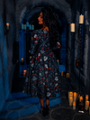 With a haunting allure, a gorgeous model graces a dimly lit dungeon while wearing the BRAM STOKER'S DRACULA Gothic Tales Swing Dress in Dracula Novelty Print by La Femme en Noir, the goth fashion brand.