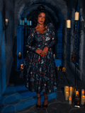 In the atmospheric gloom of a dungeon, a breathtaking model dons the BRAM STOKER'S DRACULA Gothic Tales Swing Dress in Dracula Novelty Print, a masterpiece from the goth fashion label La Femme en Noir.