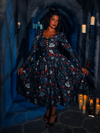 With an eerie backdrop of a dim dungeon, an alluring model showcases the BRAM STOKER'S DRACULA Gothic Tales Swing Dress in Dracula Novelty Print, a design by gothic brand La Femme en Noir.