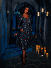 Posing in the dimly lit depths of a dungeon, a stunning model is dressed in the BRAM STOKER'S DRACULA Gothic Tales Swing Dress in Dracula Novelty Print from the goth clothing brand La Femme en Noir.