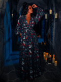 The dungeon room's candlelit ambiance sets the stage for the model to display the BRAM STOKER'S DRACULA Belladonna Maxi Dress, featuring the intriguing Dracula Novelty Print.