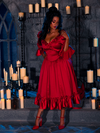 With candles spread around, a beautiful model elegantly poses in the BRAM STOKER'S DRACULA Mina Satin Bustle Dress in Blood Red in a dimly lit dungeon room.