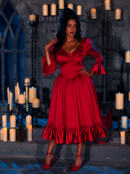 The dimly lit dungeon room sets the stage for a beautiful model to wear the BRAM STOKER'S DRACULA Mina Satin Bustle Dress in Blood Red and strike a captivating pose surrounded by candles.