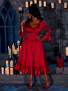 The enchanting ambiance of a dimly lit dungeon room is accentuated as a beautiful model dons the BRAM STOKER'S DRACULA Mina Satin Bustle Dress in Blood Red, surrounded by candles.