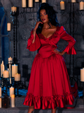 In a dimly lit dungeon room, a stunning model strikes a pose wearing the BRAM STOKER'S DRACULA Mina Satin Bustle Dress in Blood Red, surrounded by candles.