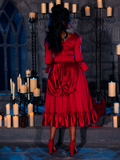 A beautiful model stands amidst candles spread around a dimly lit dungeon room, exuding grace while wearing the BRAM STOKER'S DRACULA Mina Satin Bustle Dress in Blood Red.