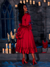 Amidst the warm candlelight in a dimly lit dungeon room, a beautiful model poses elegantly in the BRAM STOKER'S DRACULA Mina Satin Bustle Dress in Blood Red.